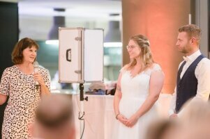 Traurednerin Susanne Schagne spricht zum Brautpaar Traurednerin Susanne Schagen hält eine emotionale Rede während einer freien Trauung, mit einem strahlenden Lächeln und liebevollen Worten, während das Brautpaar gerührt zuhört.